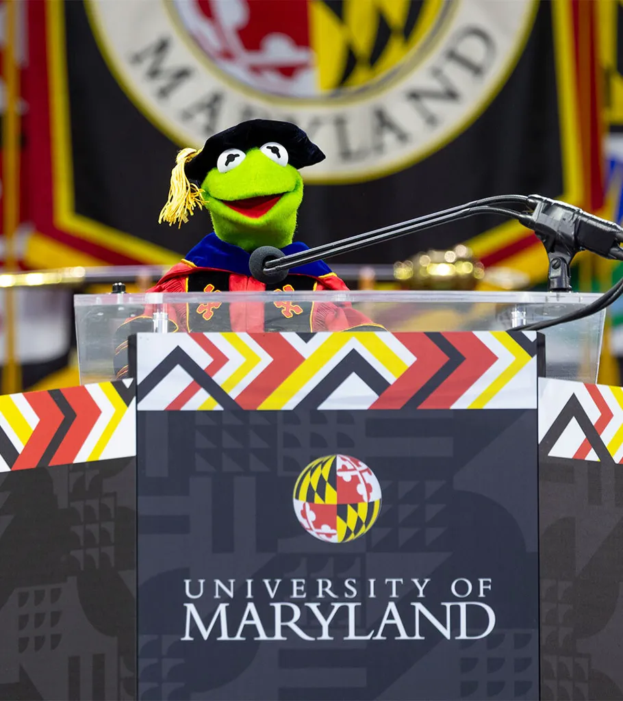 Kermit the Frog wearing academic regalia as he speaks at podium