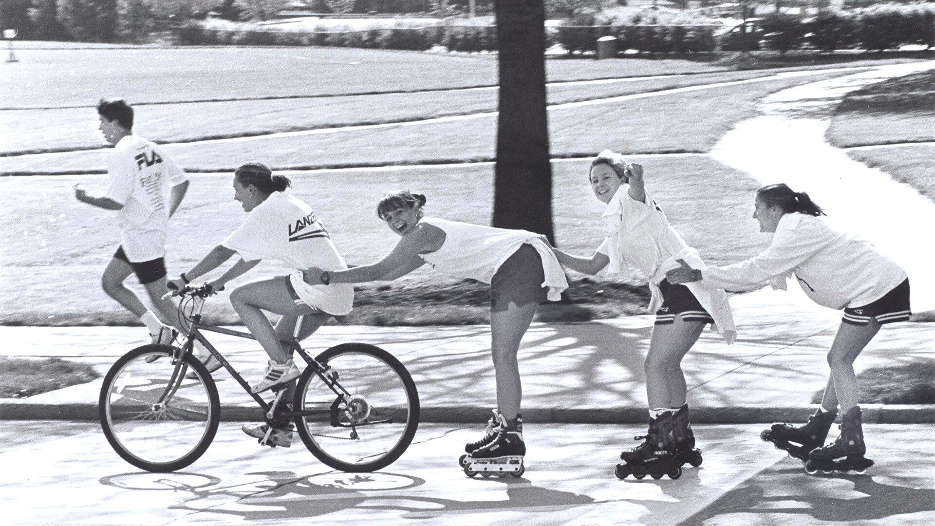 one student on bike pulls three others on roller skates, with one student running next to them